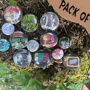 May include: A collection of 14 round pinback buttons with various designs, including text and graphics. The buttons feature phrases like "Definitely Not Dead" and "Ghost Tour." The buttons are arranged on a mossy surface.