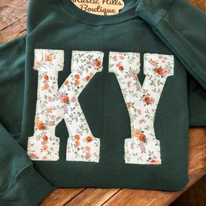 May include: A dark green sweatshirt with the letters "KY" in large white letters with a floral pattern. The sweatshirt has a tag that says "Rustic Hills Boutique".
