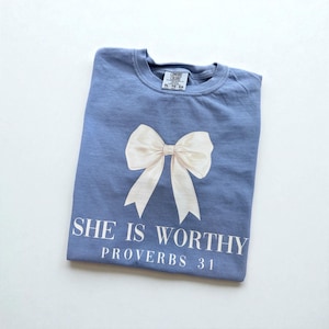 May include: A blue t-shirt with a large white bow graphic. The text "SHE IS WORTHY PROVERBS 31" is printed below the bow in white. The shirt is folded neatly on a white surface.