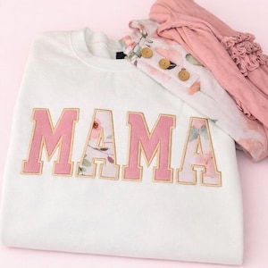 May include: White sweatshirt with the word "MAMA" in pink and floral-patterned letters. A floral-print top with wooden buttons and a pink ruffled garment are also visible, all on a pink background.