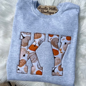 May include: A light gray sweatshirt with the letters "KY" appliqued on the front. The letters are filled with a pumpkin-themed fabric in shades of orange, brown, and cream. The sweatshirt is displayed on a white, fluffy surface.