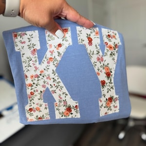 May include: A light blue sweatshirt with the letters "KY" appliqued on the front. The letters are made of a floral fabric with red, pink, and orange flowers. The edges of the letters are stitched with a contrasting thread.