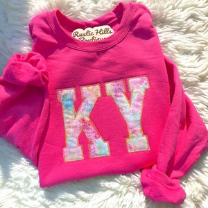 May include: Pink long-sleeve t-shirt with a tie-dye "KY" graphic. The shirt has a crew neck and is made of a soft, comfortable fabric. Rustic Hills Boutique.