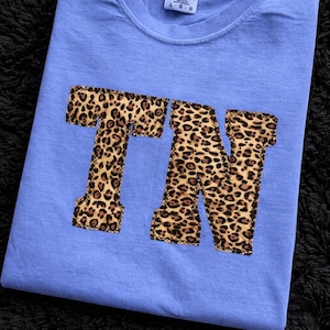 Puede incluir: Camiseta azul claro con las letras "TN" en estampado de leopardo. La camiseta está doblada sobre una superficie negra texturizada. La marca "Comfort Colors" es visible en la etiqueta.