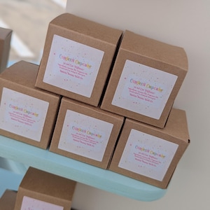 May include: A stack of brown cardboard boxes containing 10 ml car diffusers. Each box features a white label with the text Confetti Cupcake, 10 ml Car Diffuser, ingredients, and Bubble Bunny Bath Co. The labels have colorful confetti accents.