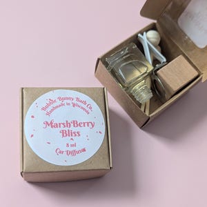 May include: A car air freshener set in a small cardboard box. The box has a round sticker with the text "Marsh Berry Bliss" and "Bubble Bunny Bath Co., Handmade in Wisconsin." The set includes a glass bottle, wooden diffuser, and accessories. The bottle contains 8 ml of liquid.