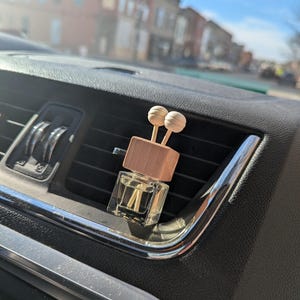 May include: A car air freshener with a clear glass bottle and a wooden top, featuring two spherical wooden accents. The air freshener is placed in a car's air vent, with a blurred cityscape visible in the background.