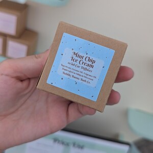 May include: A square brown cardboard box containing a 10 ml car diffuser. The label is light blue with dark speckles and features the text Mint Chip Ice Cream, 10 ml Car Diffuser, Ingredients: Coconut Oil, Fragrance Oil, Bubble Bunny Bath Co.