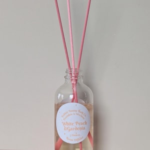 May include: A clear glass room diffuser bottle with pink reeds. The bottle contains a light-colored liquid and a label that reads "White Peach & Gardenia." The diffuser is 3 ounces.
