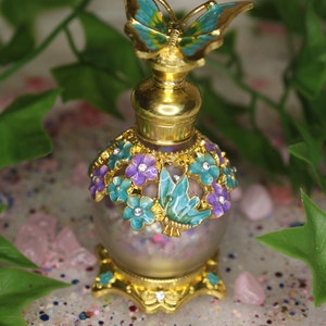 Puede incluir: Un frasco de perfume dorado con un tapón de mariposa y detalles florales. El frasco está lleno de purpurina y tiene un cuerpo de vidrio transparente.