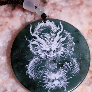 Puede incluir: Colgante redondo verde oscuro con un dise&ntilde;o detallado de drag&oacute;n blanco. El colgante est&aacute; suspendido de un cord&oacute;n negro con un cierre blanco y una peque&ntilde;a cuenta verde. El drag&oacute;n est&aacute; intrincadamente tallado, mostrando escamas y una expresi&oacute;n feroz.