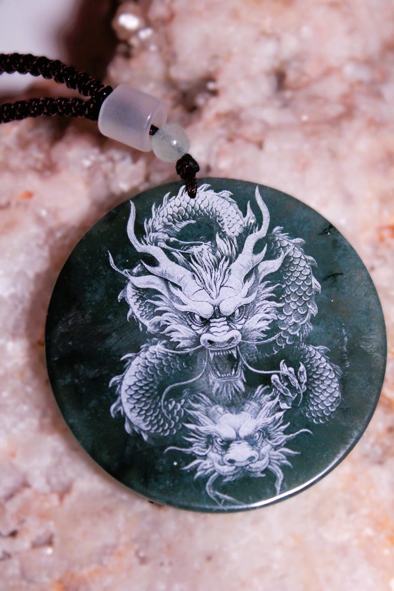 Puede incluir: Colgante redondo verde oscuro con un dise&ntilde;o detallado de drag&oacute;n blanco. El colgante est&aacute; suspendido de un cord&oacute;n negro con un cierre blanco y una peque&ntilde;a cuenta verde. El drag&oacute;n est&aacute; intrincadamente tallado, mostrando escamas y una expresi&oacute;n feroz.