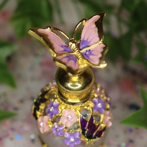 May include: A gold and purple glass bottle with a butterfly stopper. The bottle is decorated with flowers and rhinestones.