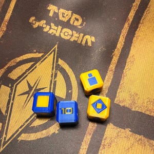 Pode incluir: Quatro dados amarelos e azuis com desenhos geométricos. Dois dados são amarelos com quadrados e losangos azuis, e dois são azuis com quadrados amarelos e o número 10. Os dados estão sobre uma superfície castanha com texto dourado e um desenho geométrico.