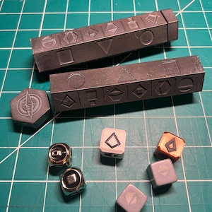 May include: A selection of game components in different shapes and colours. Two dark grey rectangular bars with geometric symbols are shown with a grey compass-shaped piece, dice, and metallic pieces with geometric designs. The pieces are on a green grid.