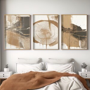 Op de afbeelding: Drie abstracte kunstprints in gouden, bruine en witte tinten. De prints zijn ingelijst in goud en hangen aan een witte muur boven een bed met een bruin dekbed.