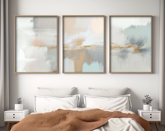May include: Three abstract paintings in gold frames, each with a different color scheme of white, gray, and blue. The paintings are hung on a white wall above a bed with a brown blanket and white pillows.
