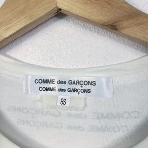 K&ouml;nnte beinhalten: Nahaufnahme des Kragens eines wei&szlig;en T-Shirts mit dem Markenetikett "COMME des GAR&Ccedil;ONS". Das Etikett ist wei&szlig; mit schwarzem Text. Darunter befindet sich ein kleineres Etikett mit der Aufschrift "SS". Das T-Shirt h&auml;ngt an einem Holzkleiderb&uuml;gel.