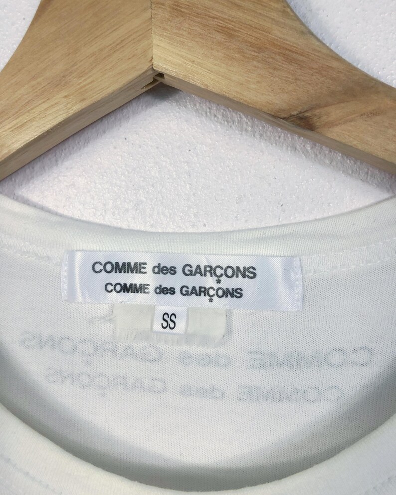 K&ouml;nnte beinhalten: Nahaufnahme des Kragens eines wei&szlig;en T-Shirts mit dem Markenetikett "COMME des GAR&Ccedil;ONS". Das Etikett ist wei&szlig; mit schwarzem Text. Darunter befindet sich ein kleineres Etikett mit der Aufschrift "SS". Das T-Shirt h&auml;ngt an einem Holzkleiderb&uuml;gel.