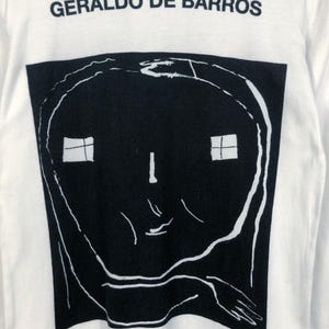K&ouml;nnte beinhalten: Wei&szlig;es Langarmshirt mit dem Text "GERALDO DE BARROS" &uuml;ber einer gro&szlig;en schwarz-wei&szlig;en Grafik. Die Grafik ist ein stilisiertes Gesicht mit zwei quadratischen Fenstern und einer Hand.