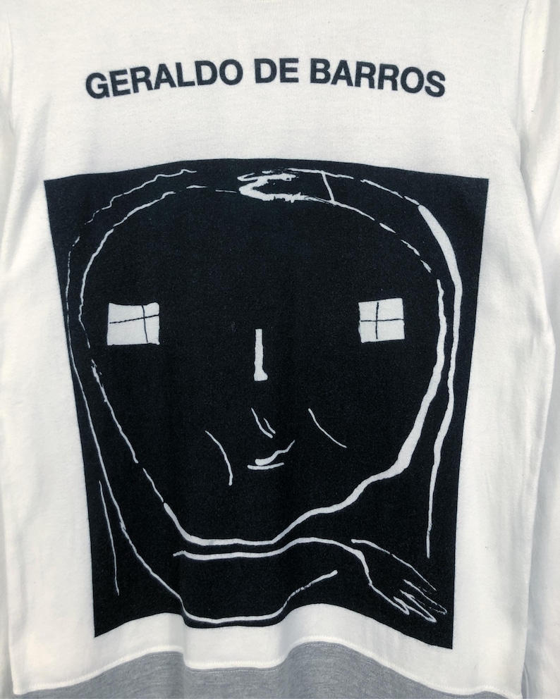K&ouml;nnte beinhalten: Wei&szlig;es Langarmshirt mit dem Text "GERALDO DE BARROS" &uuml;ber einer gro&szlig;en schwarz-wei&szlig;en Grafik. Die Grafik ist ein stilisiertes Gesicht mit zwei quadratischen Fenstern und einer Hand.