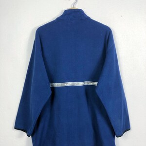 May include: A navy blue fleece pullover with a high collar and long sleeves. A white horizontal stripe with text is across the chest. The sleeves have black trim. The pullover is hanging on a metal hanger.