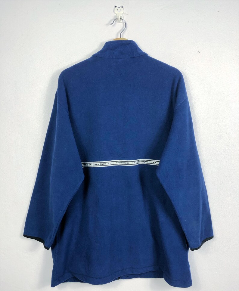 May include: A navy blue fleece pullover with a high collar and long sleeves. A white horizontal stripe with text is across the chest. The sleeves have black trim. The pullover is hanging on a metal hanger.