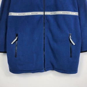 May include: A blue fleece jacket with a full-length zipper and two side pockets. The jacket features a white band across the chest with the word "courreges" printed repeatedly. Black trim accents the cuffs and hem.