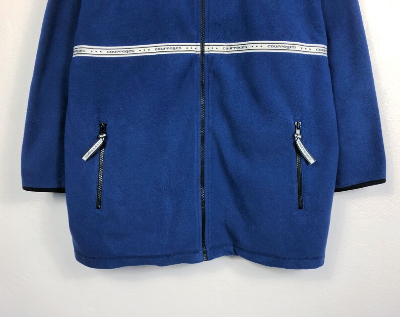 May include: A blue fleece jacket with a full-length zipper and two side pockets. The jacket features a white band across the chest with the word "courreges" printed repeatedly. Black trim accents the cuffs and hem.
