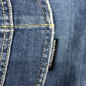 Op de afbeelding: Close-up van blauwe denimstof met gele stiksels. Een zwart label met de tekst "merci beaucoup" is zichtbaar. De afbeelding benadrukt de textuur en details van de jeans.