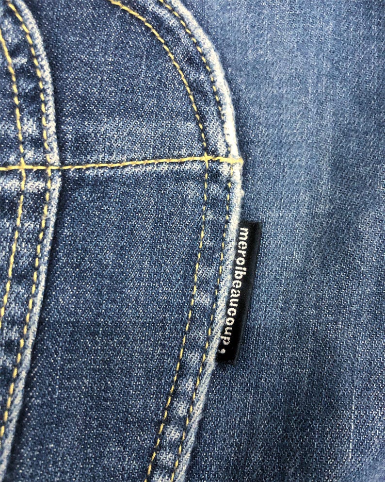 Op de afbeelding: Close-up van blauwe denimstof met gele stiksels. Een zwart label met de tekst "merci beaucoup" is zichtbaar. De afbeelding benadrukt de textuur en details van de jeans.