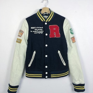 May include: A navy blue and white varsity jacket with yellow and white stripes on the cuffs and bottom. The jacket has a large red "R" on the left side of the chest and the text "ROXY is the brand that expresses the coastal & mountain casual lifestyle" on the right side of the chest.