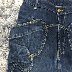 Op de afbeelding: Close-up van een donkerblauwe denim jeans met een uniek zakontwerp. De jeans heeft een knoopsluiting en gele stiksels. De zak heeft een kenmerkende gebogen vorm. De stof lijkt een mediumgewicht denim te zijn.