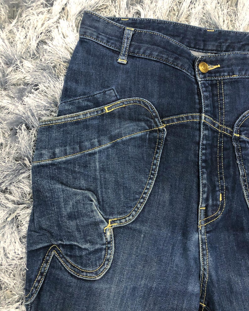 Op de afbeelding: Close-up van een donkerblauwe denim jeans met een uniek zakontwerp. De jeans heeft een knoopsluiting en gele stiksels. De zak heeft een kenmerkende gebogen vorm. De stof lijkt een mediumgewicht denim te zijn.