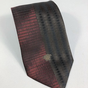 Puede incluir: Una corbata de dos tonos con un diseño burdeos y negro. La sección burdeos tiene un patrón texturizado, mientras que la sección negra tiene un patrón de rayas. Un emblema dorado está cerca de la parte inferior.