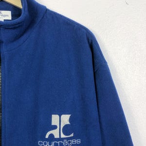 May include: A navy blue fleece jacket with a high collar and a full zipper. The jacket features a white logo and the word "courr&egrave;ges" in a matching font. The jacket is made of a soft, warm material.