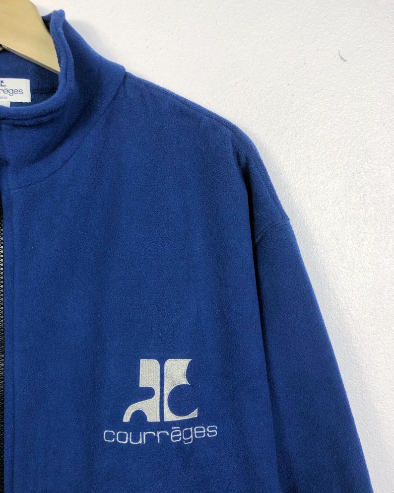 May include: A navy blue fleece jacket with a high collar and a full zipper. The jacket features a white logo and the word "courr&egrave;ges" in a matching font. The jacket is made of a soft, warm material.