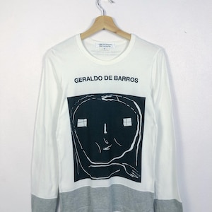 K&ouml;nnte beinhalten: Wei&szlig;es Langarmshirt mit einem schwarz-wei&szlig;en grafischen Gesichtsdesign. Das Shirt tr&auml;gt den Text "GERALDO DE BARROS" in Schwarz. Der untere Saum und die &Auml;rmelb&uuml;ndchen sind grau.