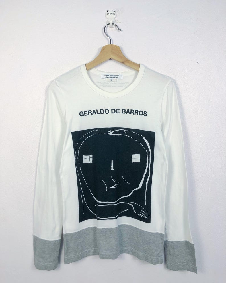 K&ouml;nnte beinhalten: Wei&szlig;es Langarmshirt mit einem schwarz-wei&szlig;en grafischen Gesichtsdesign. Das Shirt tr&auml;gt den Text "GERALDO DE BARROS" in Schwarz. Der untere Saum und die &Auml;rmelb&uuml;ndchen sind grau.