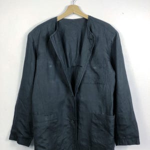 Pode incluir: Um blazer azul marinho escuro com fecho de um botão e dois bolsos frontais. O blazer é feito de um tecido leve e tem um ajuste descontraído. Está pendurado num cabide de madeira contra um fundo branco.