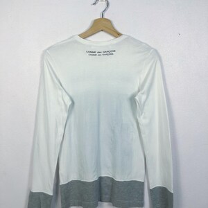 K&ouml;nnte beinhalten: Wei&szlig;es Langarmshirt mit grauen B&uuml;ndchen und Saum. Der Text "COMME des GAR&Ccedil;ONS" ist in Schwarz auf dem oberen R&uuml;cken aufgedruckt. Das Shirt h&auml;ngt an einem Holzkleiderb&uuml;gel.