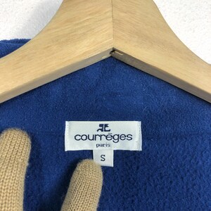May include: Close-up of a blue garment with a white label that reads "courr&egrave;ges paris" and a size "S". The garment is hanging on a wooden hanger.