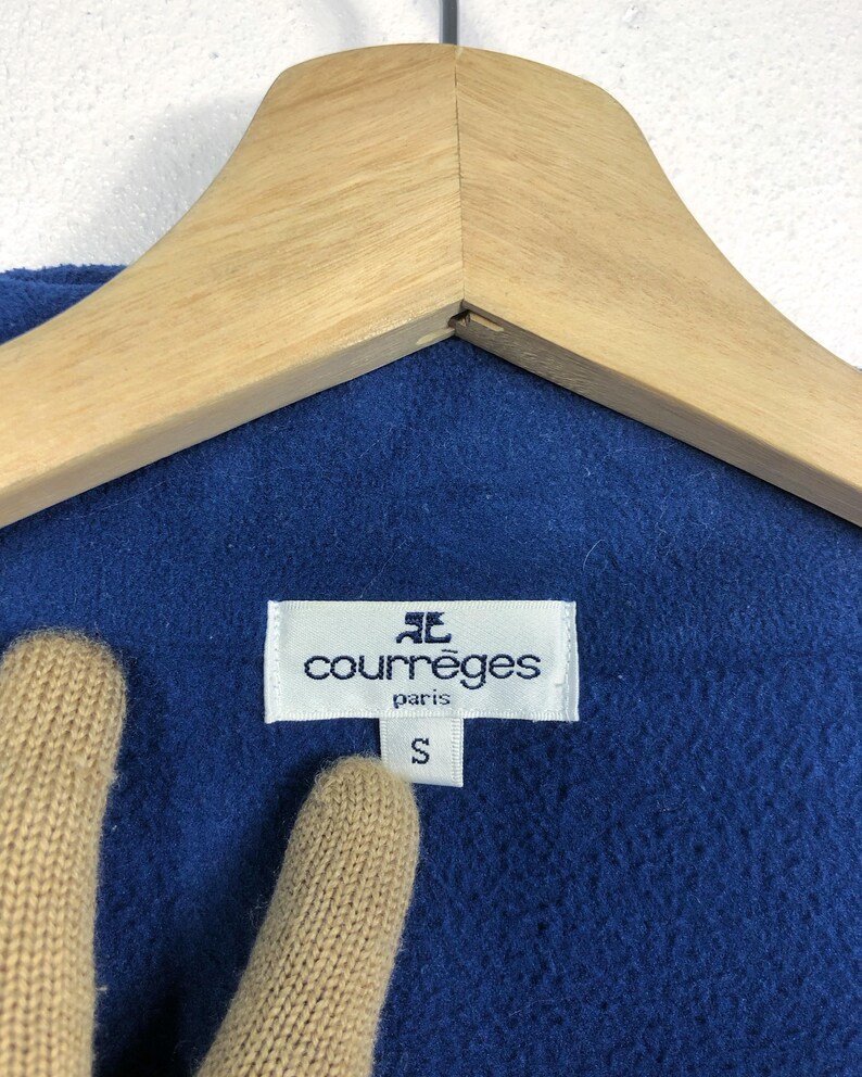 May include: Close-up of a blue garment with a white label that reads "courr&egrave;ges paris" and a size "S". The garment is hanging on a wooden hanger.