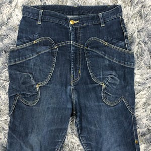 Op de afbeelding: Donkerblauwe spijkerbroek met unieke zakontwerpen en een gouden knoopsluiting. De jeans hebben kenmerkende gebogen zakvormen en decoratieve stiksels, wat een stijlvolle en moderne uitstraling geeft. De denim heeft een licht vervaagde uitstraling.