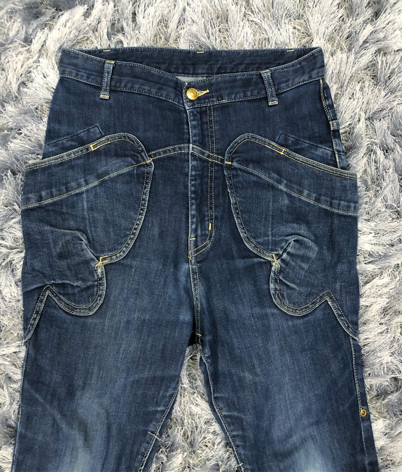 Op de afbeelding: Donkerblauwe spijkerbroek met unieke zakontwerpen en een gouden knoopsluiting. De jeans hebben kenmerkende gebogen zakvormen en decoratieve stiksels, wat een stijlvolle en moderne uitstraling geeft. De denim heeft een licht vervaagde uitstraling.