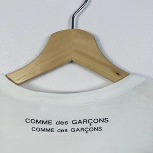 K&ouml;nnte beinhalten: Wei&szlig;es T-Shirt mit dem schwarzen Aufdruck "COMME des GAR&Ccedil;ONS". Das Kleidungsst&uuml;ck h&auml;ngt an einem Holzkleiderb&uuml;gel mit Metallhaken. Der Kragen ist sichtbar, vor einer schlichten wei&szlig;en Wand.