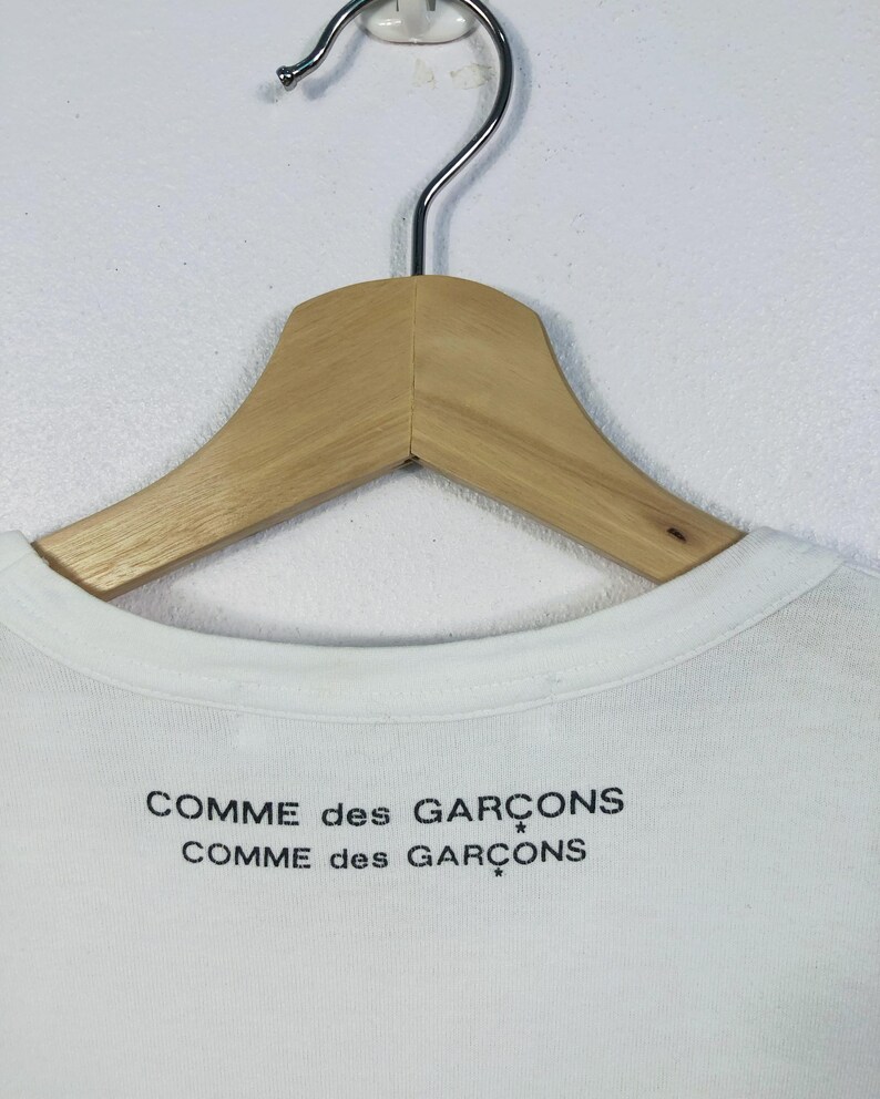 K&ouml;nnte beinhalten: Wei&szlig;es T-Shirt mit dem schwarzen Aufdruck "COMME des GAR&Ccedil;ONS". Das Kleidungsst&uuml;ck h&auml;ngt an einem Holzkleiderb&uuml;gel mit Metallhaken. Der Kragen ist sichtbar, vor einer schlichten wei&szlig;en Wand.