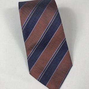 Puede incluir: Una corbata a rayas diagonales en tonos óxido, azul marino y morado claro. La corbata tiene un aspecto texturizado y probablemente está hecha de seda o un material similar. Las rayas están espaciadas uniformemente.