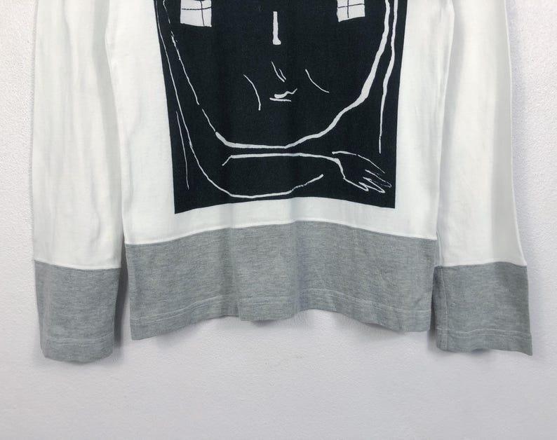 K&ouml;nnte beinhalten: Wei&szlig;es Langarmshirt mit grauem Saum und B&uuml;ndchen. Das Shirt hat eine gro&szlig;e schwarz-wei&szlig;e Grafik eines Gesichts mit Arm und Hand. Das Design ist auf der Vorderseite zentriert.