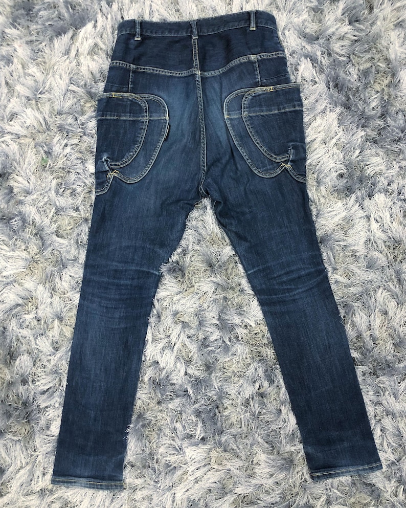Op de afbeelding: Donkerblauwe denim jeans met unieke zakdetails. De jeans heeft grote, gebogen zakken op de achterkant en een hoge taille. De denim heeft subtiele vervaging en slijtage, wat het een vintage look geeft. De jeans ligt plat op een getextureerd oppervlak.